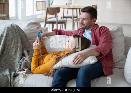 Fröhliche Frau, die auf den Knien des Mannes liegt, Smartphone hält, lächelt, während sie mit Verwandten Video hat Stockfoto