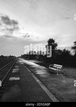 Schwarz-weiß-Blick auf einen einsamen Bahnsteig mit einer Bank und Schienen, die am Horizont verschwinden Stockfoto