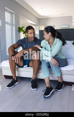 Afroamerikaner und asiatische Frau sitzen auf dem Bett, lächeln und reden zu Hause Stockfoto