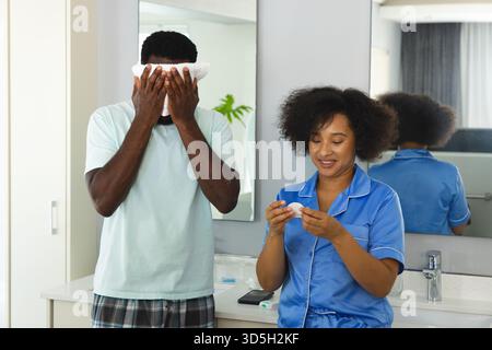 Junges Paar im modernen Badezimmer, Mann, der ein Handtuch in Gesicht hält, während die Frau das Produkt auf dem Pad aufträgt Stockfoto