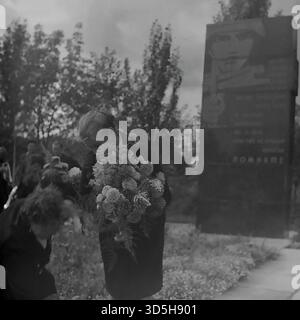 UdSSR, Ukrainische SSR, Slawjansk, 1975. Ein zutiefst bewegender, ehrlicher Schuss aus der Zeremonie zum 30. Siegtag an einer Gedenkstätte im Stadtteil Keram. Ein älterer Mensch, der sich vor Trauer verbeugt, hält einen großen Strauß Chrysanthemen, bereit, sie zu legen. Im Hintergrund ist die große Gedenkstele mit dem Wort „Pomnim“ (wir erinnern uns) unscharf sichtbar. Dieses archivierte Schwarzweiß-Foto zeigt einen persönlichen und feierlichen Akt der Erinnerung eines Einheimischen während eines großen sowjetischen Feiertags. Stockfoto