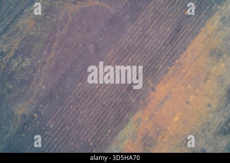 Luftaufnahme von oben nach unten bearbeiteter landwirtschaftlicher Böden mit starken diagonalen Linearmustern. Gedämpfte, kalte Violettgrau-Töne stehen im Kontrast zu warmen, rostigen Patches. Konzept für Landwirtschaft, Klima. Stockfoto