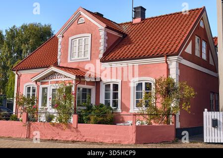 AHUS, Schweden; 22. September 2025: Bezauberndes rosafarbenes Ziegelhaus mit weißer Zierleiste und rotem Ziegeldach entlang der Hauptstraße, dargestellt in warmem Sonnenlicht Stockfoto