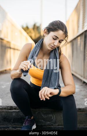 Frau ruht aus, überprüft Aktivitätstracker nach dem Training, trägt Sportbekleidung mit Handtuch Stockfoto