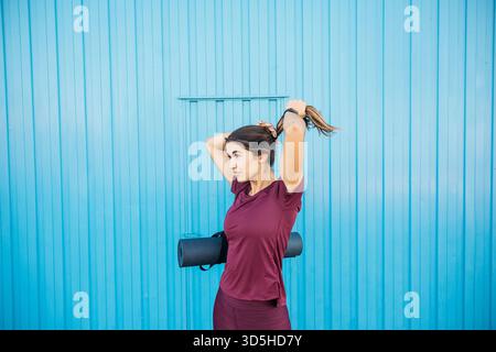 Frau, die ihr Haar bindet, eine Yogamatte hält, sich auf ihre Fitness vorbereitet Stockfoto