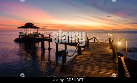 Entspannen Sie sich auf einem hölzernen Pier, während die Sonne über dem ruhigen Wasser der Insel Koh Kood untergeht. Der Himmel ist mit lebhaften Orange- und Rosa-Tönen gefüllt und bietet eine wunderschöne Kulisse für einen abendlichen Rückzugsort in Thailand Stockfoto