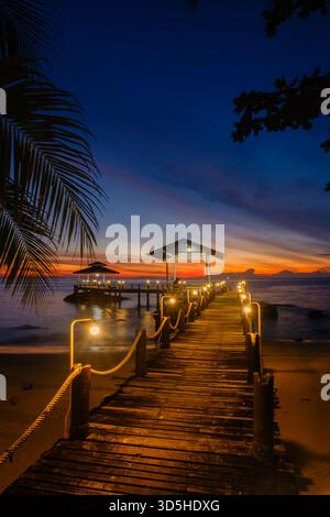 Ein atemberaubender Sonnenuntergang wirft lebendige Farben über den Horizont auf Koh Kood Island. Der hölzerne Pier mit warmen Lichtern schafft eine ruhige Atmosphäre, die sich perfekt für Abendspaziergänge am Strand eignet. Stockfoto