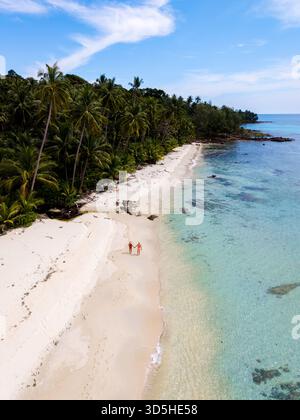 Zwei Strandgänger genießen einen ruhigen Spaziergang entlang des unberührten weißen Sandes von Koh Kood Island. Das türkisfarbene Wasser schimmert unter einem klaren blauen Himmel, umgeben von üppigen Palmen und lebhaftem Grün Thailands Stockfoto