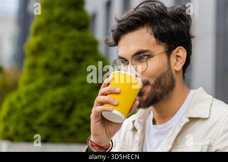 Glücklicher junger indischer Mann, der morgens Kaffee, heißes Getränk genießt und draußen lächelt. Entspannen, eine Pause einlegen. Ein arabischer Hindu-Typ in der Innenstadt, trinkt Kaffee zum Mitnehmen. Stadtleben draußen Stockfoto