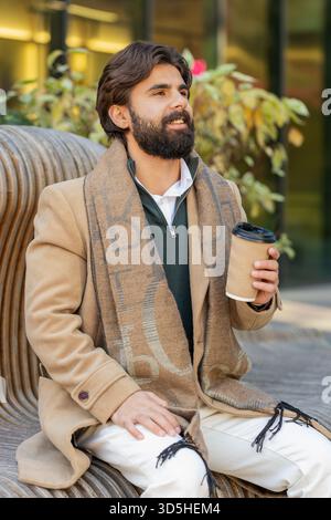 Glücklicher junger indischer Mann, der morgens Kaffee, ein heißes Getränk genießt und draußen lächelt. Entspannen, eine Pause einlegen. Der libanesische Kerl sitzt auf den Stadtmitten Stockfoto