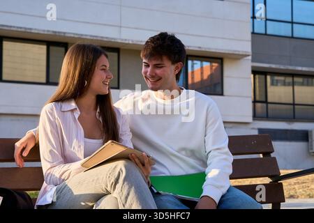 Zwei fröhliche junge Studenten sitzen auf einer Universitätsbank, teilen sich einen Moment und schauen sich ein Tablet an Stockfoto