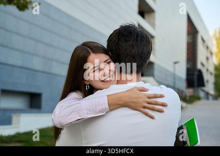 Eine junge Frau lächelt, während sie draußen einen Mann umarmt. Die Schüler teilen sich einen liebevollen Moment nach dem Unterricht Stockfoto