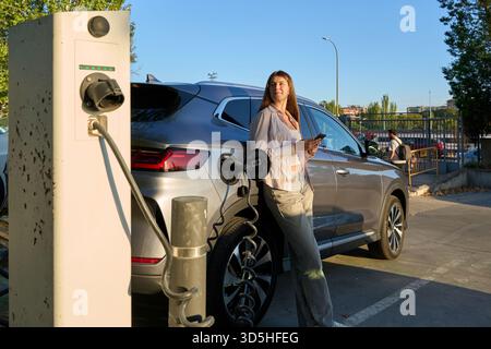 Frau, die neben dem Aufladen eines Elektroautos steht und Smartphone hält. Konzept für umweltfreundliches Reisen, erneuerbare Energien und modernen Lebensstil Stockfoto