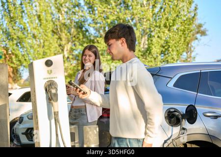 Junge Paare, die ihr Smartphone benutzen, während sie ein Elektrofahrzeug an einer öffentlichen Station aufladen, um einen nachhaltigen Transport zu fördern Stockfoto