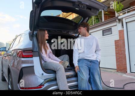 Junge Paare, die ihr Gepäck in einen elektrischen suv packen, während sie darauf warten, dass das Fahrzeug vor der Fahrt aufgeladen wird Stockfoto