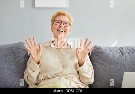 Ältere Frau drückt Freude aus, wenn sie bequem auf einem weichen Sofa in ihrem Wohnzimmer sitzt. Stockfoto