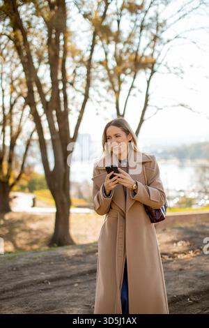 Eine Frau mit modischem Mantel steht in einem Park mit Herbstbäumen. Sie lächelt, während sie ihr Handy überprüft und die warme Sonne und das lebhafte sce genießt Stockfoto
