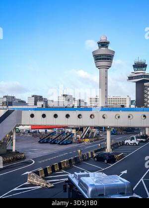 Amsterdam, Niederlande - 14. November 2023: Asphalt des Flughafenkontrollturms Schiphol mit ikonischem Kontrollturm, Terminal-Jetbrücke, Bodenunterstützungsausrüstung, Fahrzeugen, blauem Himmel. Reisen, Luftfahrt, Transport, Logistik, globaler Tourismus, Geschäftsreise Stockfoto