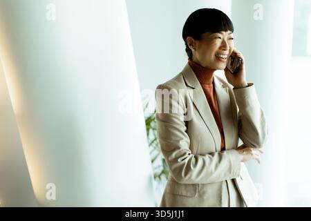 Eine professionelle Frau lächelt, während sie am Telefon spricht. Sie steht selbstbewusst in einem schlanken, gut beleuchteten Büro. Stockfoto