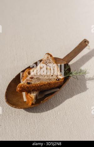 Zwei Scheiben handwerkliches getrocknetes Fruchtbrot auf rustikalem Holzbrett, roher Leinentischdecke, Rosmarin. Traditionelle Bäckerei, Brot, rustikal, authentisch Stockfoto