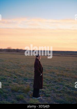 Porträt in voller Länge, eine junge Frau, die allein auf leerem Feld steht und langes braunes Fell trägt, während der bunten Sonnenuntergang oder Sonnenaufgang aussehende Kamera. Stockfoto