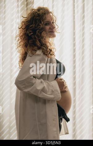 Die werdende Frau steht im Profil, lächelt, während sie sanft ihren Bauch berührt, ein weißes Hemd und dunkle Kleidung trägt, mit weicher Innenbeleuchtung. Stockfoto