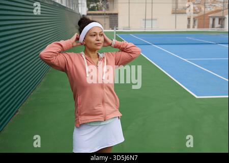 Eine Frau mit pfirsichfarbenem Hoodie und Stirnband steht mit den Händen hinter dem Kopf auf einem Tennisplatz, ruhig und konzentriert, bereit für Outdoor-Training. Stockfoto