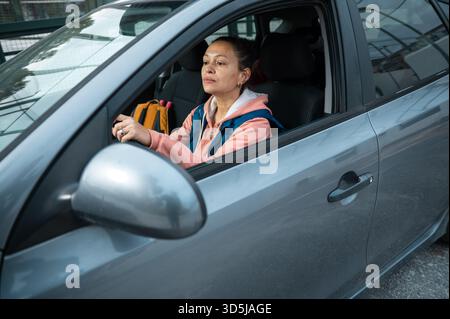 Eine Frau fährt ein graues Auto in einer urbanen Umgebung, trägt einen rosafarbenen Hoodie und eine blaue Weste. Sie blickt mit ruhiger Konzentration nach vorne, geeignet für Lifestyle und Transp Stockfoto