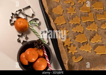 Ungebackene Lebkuchenkekse ruhen auf einem Backblech mit Backpapier, einer Schüssel mit Mandarinen und Zuckerstangen aus Metall. Bereit für Weihnachten. Stockfoto