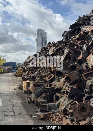 Motorblöcke und andere Metallteile stapeln sich hoch auf einem Schrottplatz, im Hintergrund erhebt sich eine Skyline der Stadt Stockfoto
