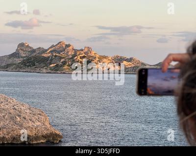 Person, die ein Smartphone hält und einen wunderschönen Sonnenuntergang über der felsigen Insel und dem Meer aufnimmt Stockfoto