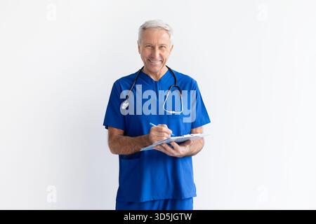 Lächelnder älterer männlicher Arzt in blauen Schutten mit Klemmbrett auf weißem Hintergrund Stockfoto