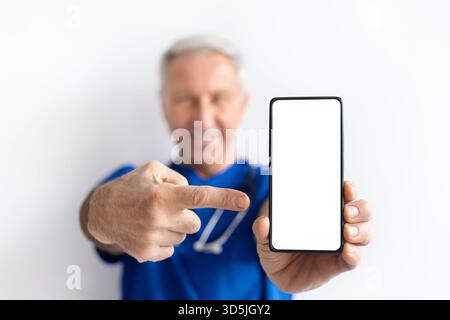 Leitender männlicher Arzt in blauer Uniform, der mit leerem Bildschirm auf das Smartphone zeigt Stockfoto
