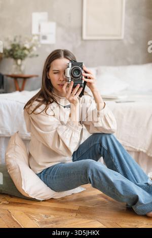 Mittelgroße Frau in Jeans, die auf dem Boden in ihrem Schlafzimmer sitzt und ein Foto mit ihrer Kamera macht Stockfoto