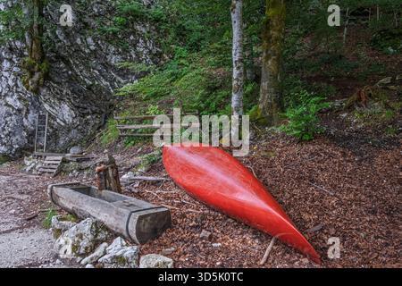 Rotes Kanu, das kopfüber in einer Waldlichtung neben rustikalen Holzstrukturen und felsigem Gelände ruht, friedliche Naturszene im Freien. Stockfoto