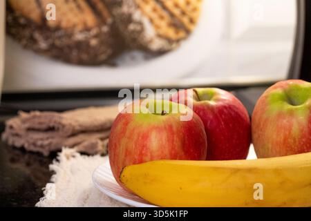 Warm beleuchtete Küchenszene mit drei Äpfeln und einer Banane auf einem weißen Teller, vor einem Hintergrund mit Lampe und Waffeln Stockfoto