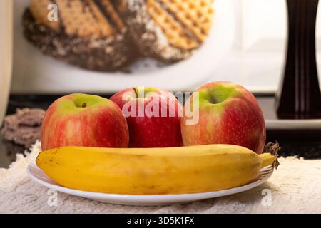Warm beleuchtete Küchenszene mit drei Äpfeln und einer Banane auf einem weißen Teller, vor einem Hintergrund mit Lampe und Waffeln Stockfoto