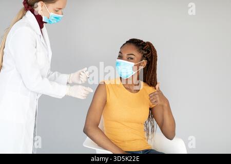 Junge Schwarze Frau erhält COVID-Impfstoff, während sie die Genehmigung mit der Geste „Daumen hoch“ zeigt Stockfoto