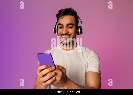 Junger arabischer Mann, der Musik und Apps auf dem Smartphone unter Neonlicht genießt Stockfoto