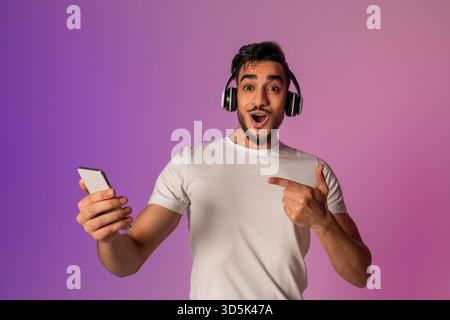Der überraschte junge arabische Mann reagiert auf die coole Mobile App, während er Kopfhörer in Neonfarben trägt Stockfoto