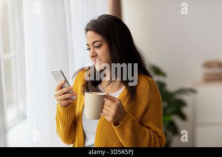 Die junge arabische Dame genießt Kaffee, während sie auf dem Smartphone in der Nähe des Fensters in gemütlicher Atmosphäre surft Stockfoto