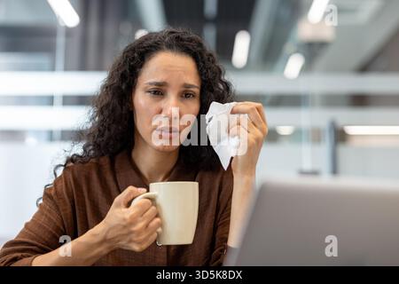 Junge Geschäftsfrau mit lockigem Haar, die ein Taschentuch und eine Kaffeetasse hält, die traurig und verärgert aussieht, während sie emotionalen Stress oder Burnout an ihrem Schreibtisch erlebt Stockfoto