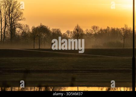 Silhouette einer einsamen Person, die während eines nebligen goldenen Sonnenaufgangs mit Wasserspiegelung an einem Flussdamm entlang geht Stockfoto