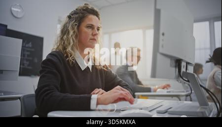Lockhaarige Frau besucht den Coding Workshop, schaut auf den Bildschirm und lächelt warm. Konzept kreatives Lernen, positive Bildungsatmosphäre, Sand Digital Empowerment im modernen Klassenzimmer. Hochformat. Stockfoto