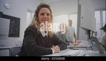 Lockhaarige Frau besucht den Coding Workshop, schaut auf den Bildschirm und lächelt warm. Konzept kreatives Lernen, positive Bildungsatmosphäre, Sand Digital Empowerment im modernen Klassenzimmer. Hochformat. Stockfoto