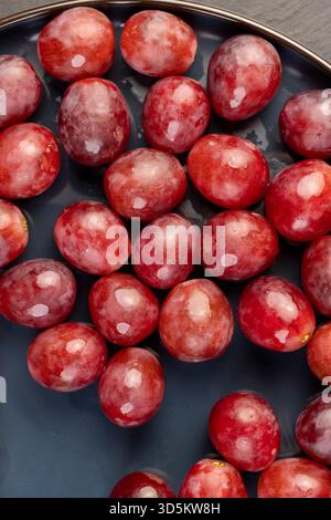 Gerissene rote Trauben in Tropfen Wasser auf einer blauen Platte, nass reife und köstliche rote Trauben auf einem Teller auf einem schwarzen Schieferbrett Stockfoto