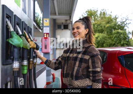 Frau lächelt und trägt einen Schutzhandschuh, während sie eine Kraftstoffpumpe auswählt, um ihr rotes Auto an einer Tankstelle aufzutanken, wobei sie die täglichen Aufgaben und die Fahrzeugeigenschaften vorstellt Stockfoto