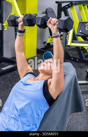 Eine Frau mittleren Alters, die mit Hanteln auf einer schrägen Bank tragend, mit einer Mütze und blauer Sportbekleidung tragend. Stockfoto