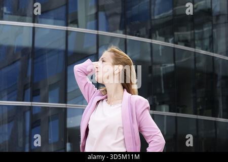 Junge schöne blonde Frau mit langen Haaren in rosa eleganten Kleidern steht in der Nähe eines modernen Gebäudes aus Glas und Stahl. Selektiver Fokus Stockfoto