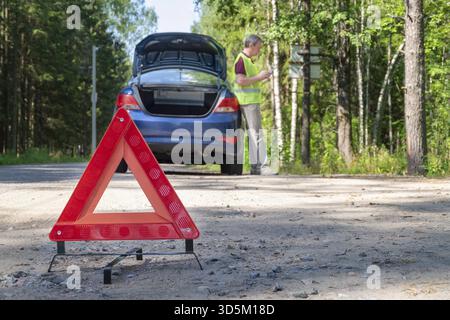 Nahaufnahme eines tragbaren reflektierenden roten Warnschilds an der Seite einer Landstraße in der Nähe eines Autos. Der Fahrer am Telefon ruft technische AS an Stockfoto